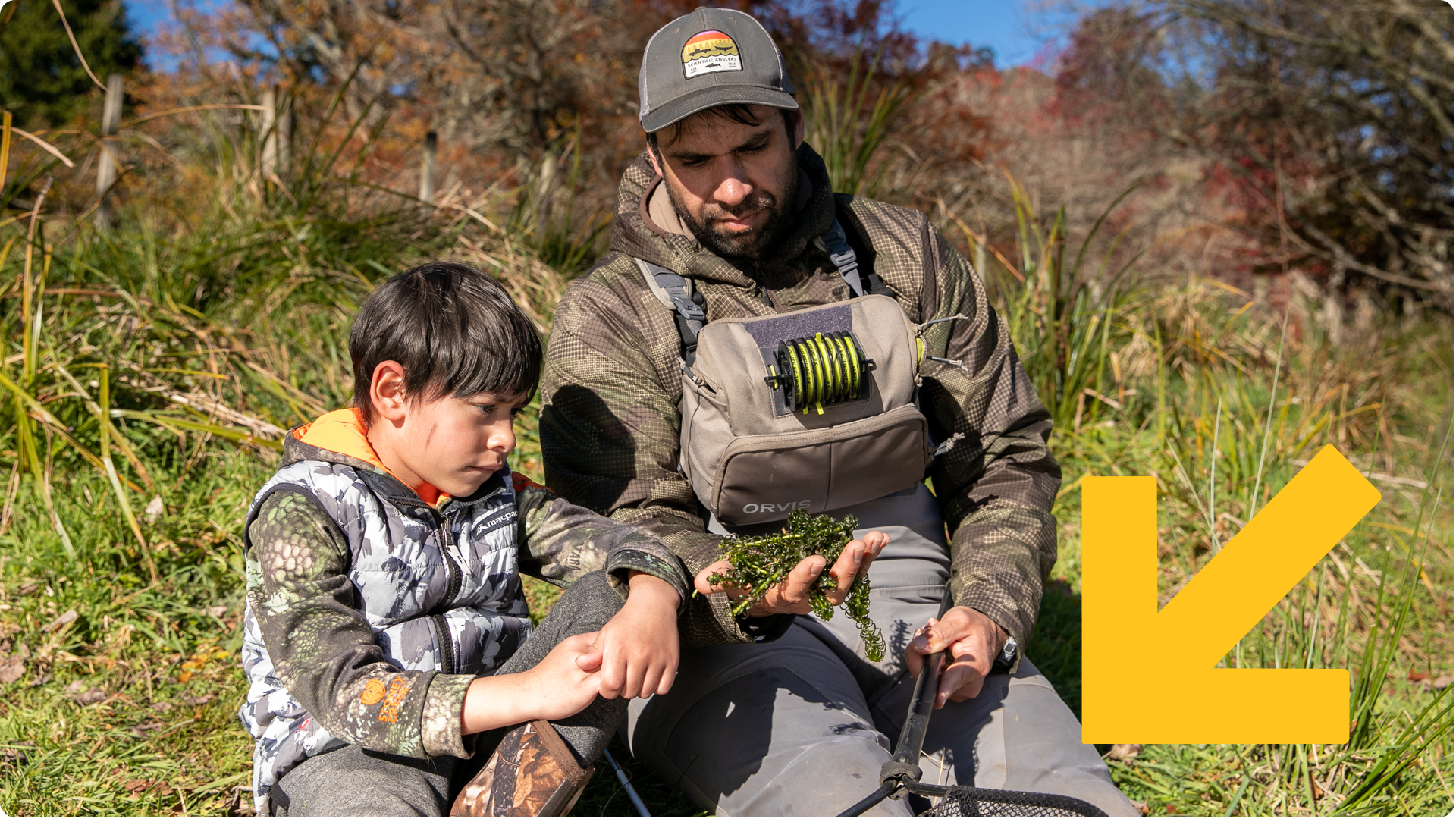  Angler showing plant material from his gear to a young boy riverside.