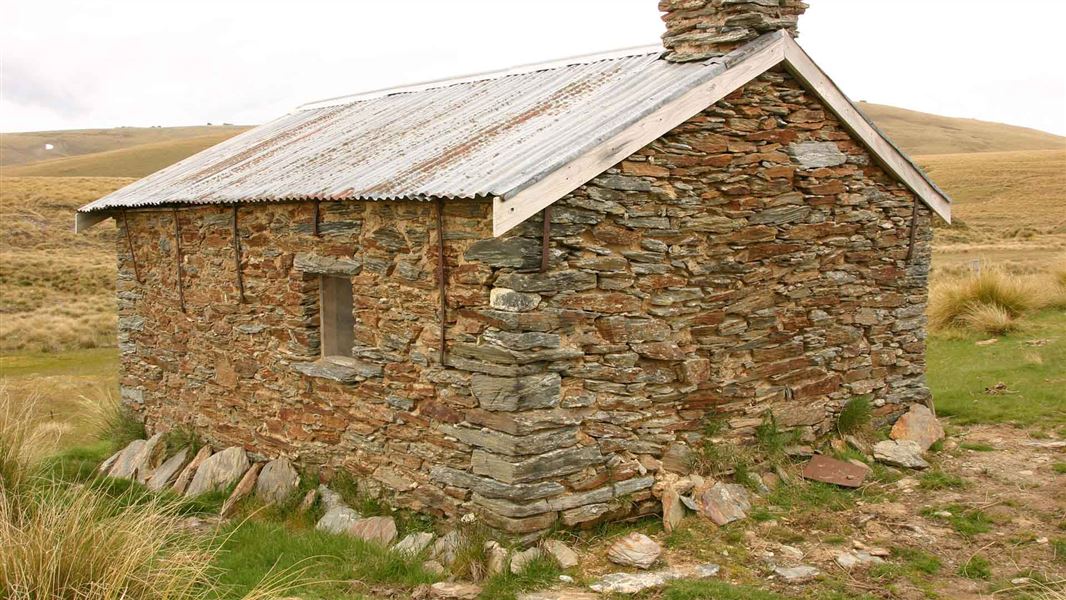 A slat rock hut amidst some short tussock. Hills roll in the background covered in tussock.