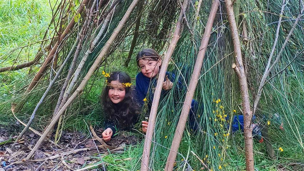 Two children laying down under a conical shape of branches to look like a hut.