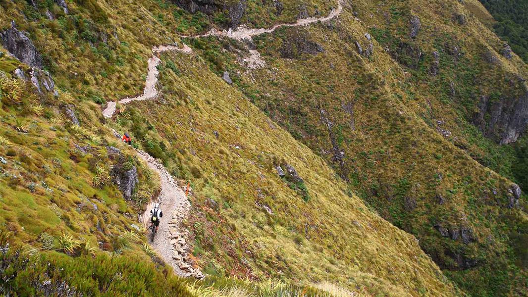 A windy track leads alongside a hilly area. Mountain bikers are riding on it