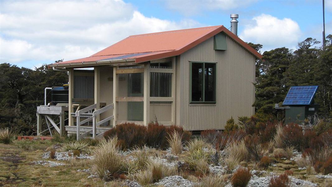 Iron clad beige DOC hut with terracotta roof and small balcony with tussocks in the foreground and trees and blue sky behind. 
