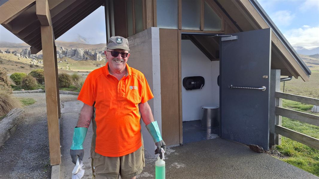 Smiling man standing outside a toilet block holding some cleaning gear.