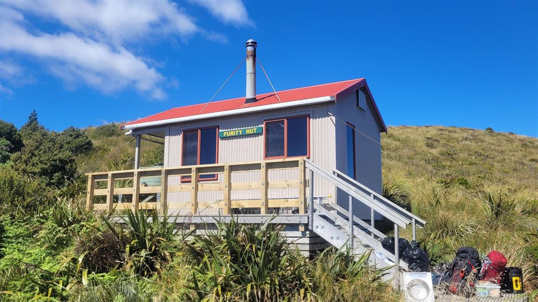 View of Purity Hut on a sunny day with blue sky. Some hiking bags are on the ground out the front of the hut.