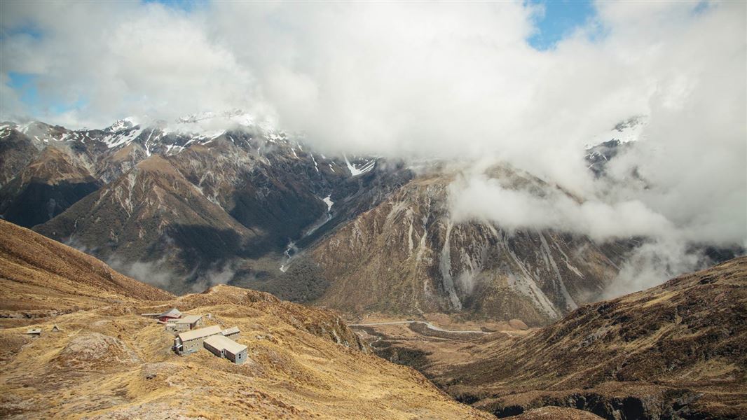 A high view over a clouded mountain valley, with three buildings in the foreground.