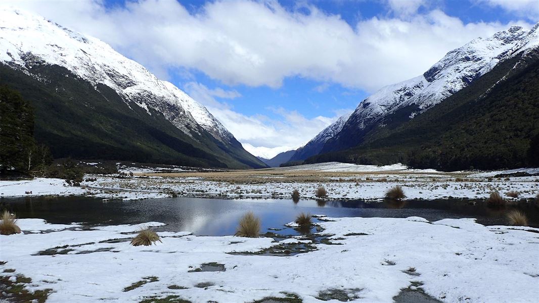 Snowy ground over a body of water with tussoclk grass coming through and snowy mountains in the background. 