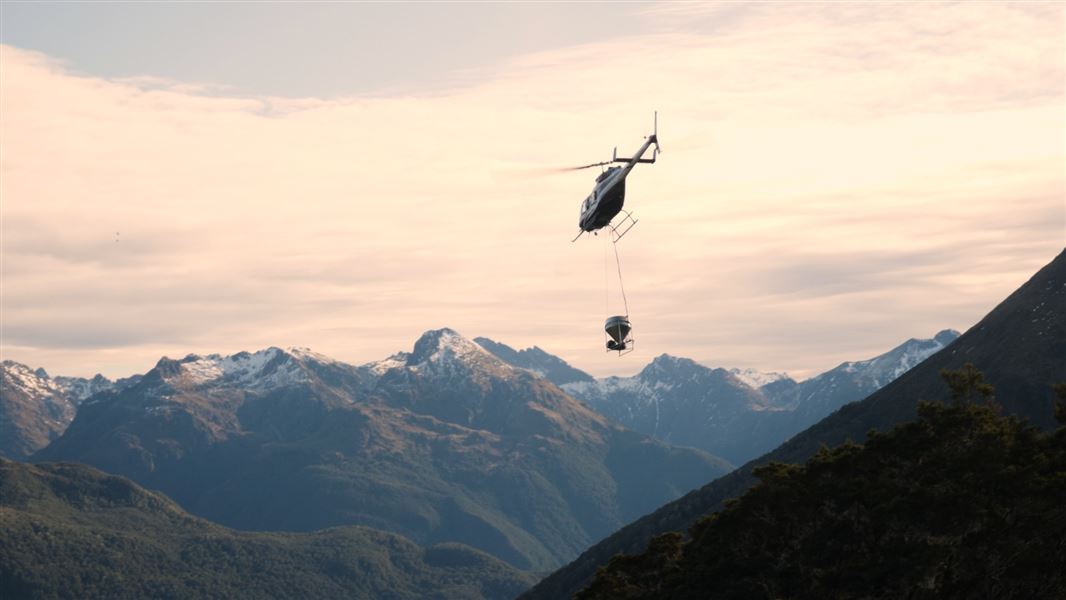 A helicopter flying over a mountain range with a drop load of 1080 biodegradable bait pellets.