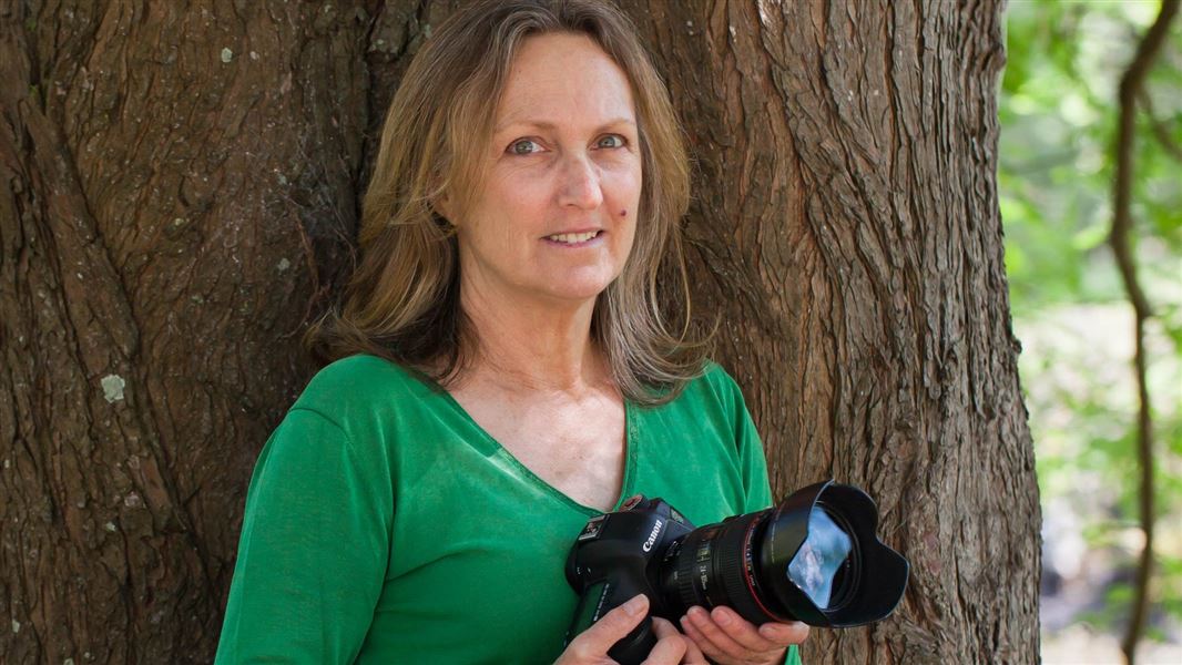 A woman leans against a tree holding photography equipment