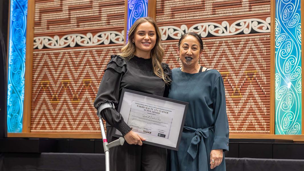Two people stand together smiling, one holds a framed diploma.
