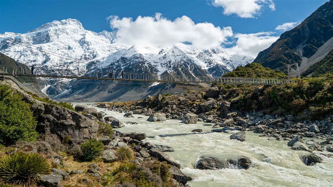 Suspension bridge crossing a rocky glacial river, with blue sky and mountains in the background