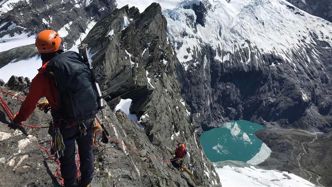 Two people in protective gear climb a mountain cliff using ropes.