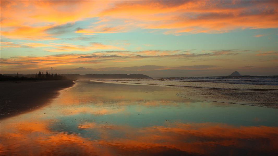 A cloudy blue sky with tinted peach clouds by setting sun, all reflected on wet sand of the beach below.