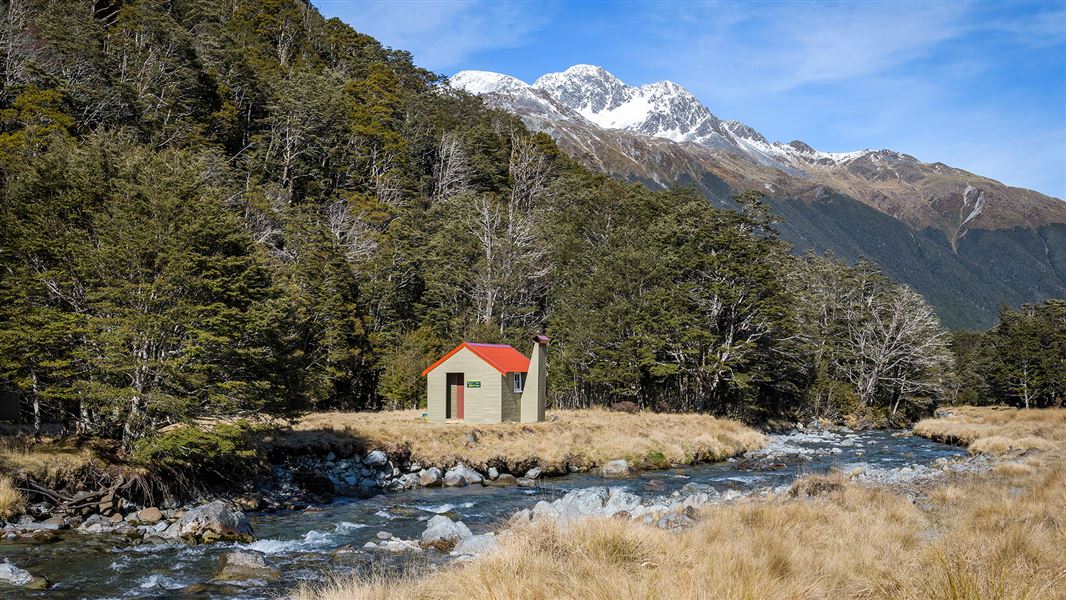 Small building by river and trees with snowy hill in background.