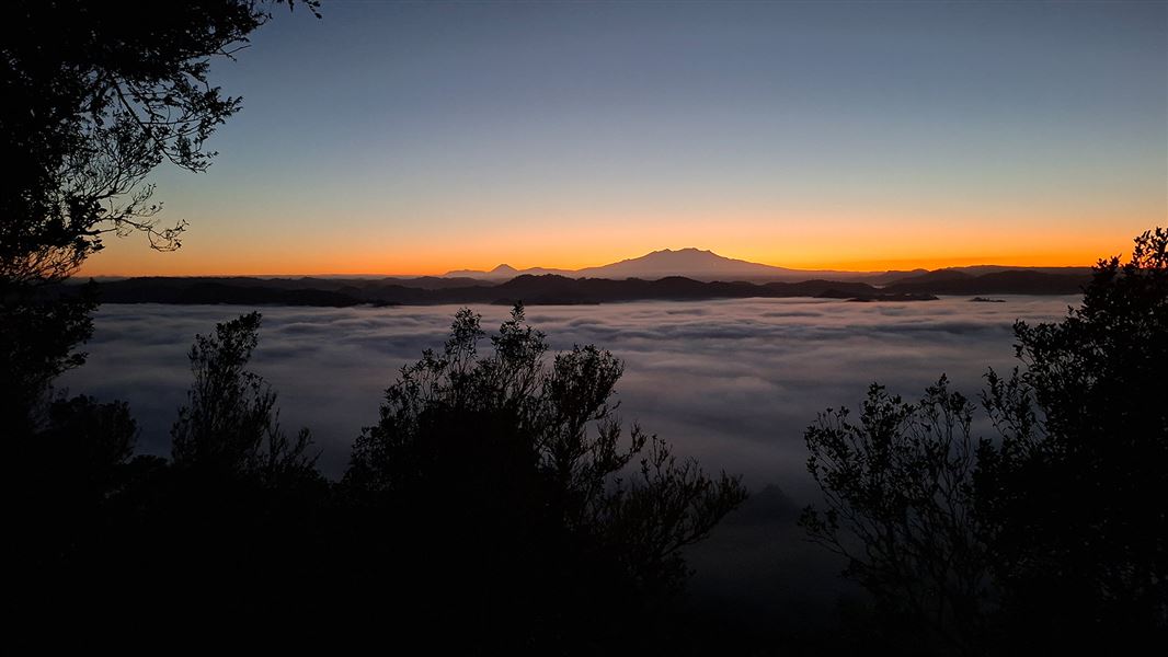 A misty floor of clouds with mountain rangge beyond covered in an orange glow at sunrise. 