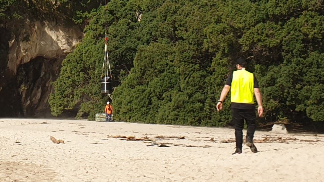 A large drum lifted by rope with two DOC staff standing nearby