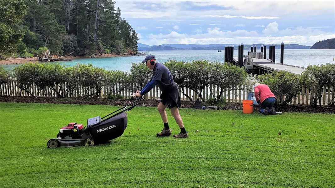 Person mowing lawns and a person weeding with a wharf in the background.