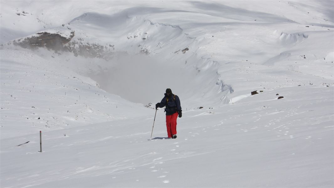 A lone tramper walks across heavy snow towards a mountain peak.