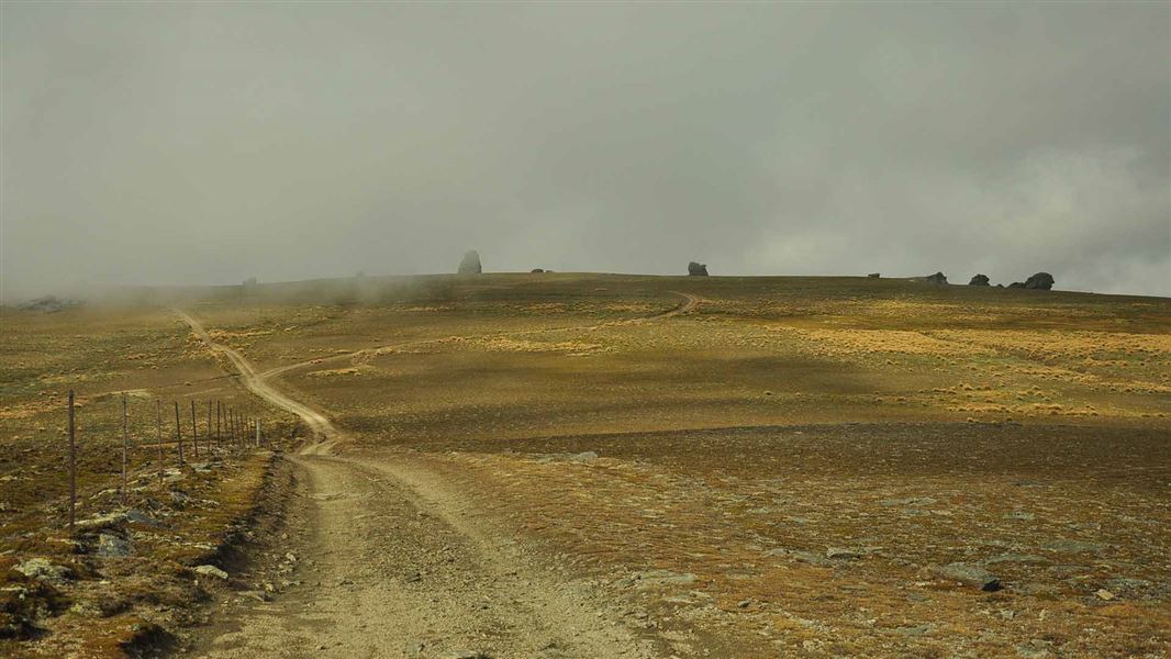 A scraggy landscape with jutting large rocks on a far away ridge. Two tracks close together created by a vehicle can be seen extending from where the photos has been taken out and over the far away ridge.