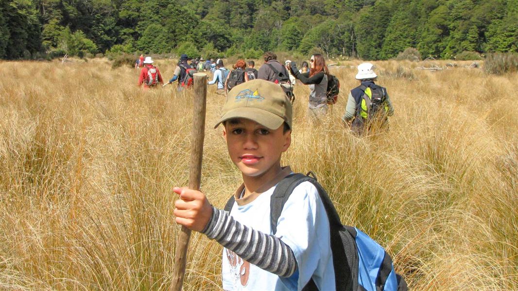 Child with a walking stick standing in a golden field of tall grass. A group of people walk through it the behind him. 