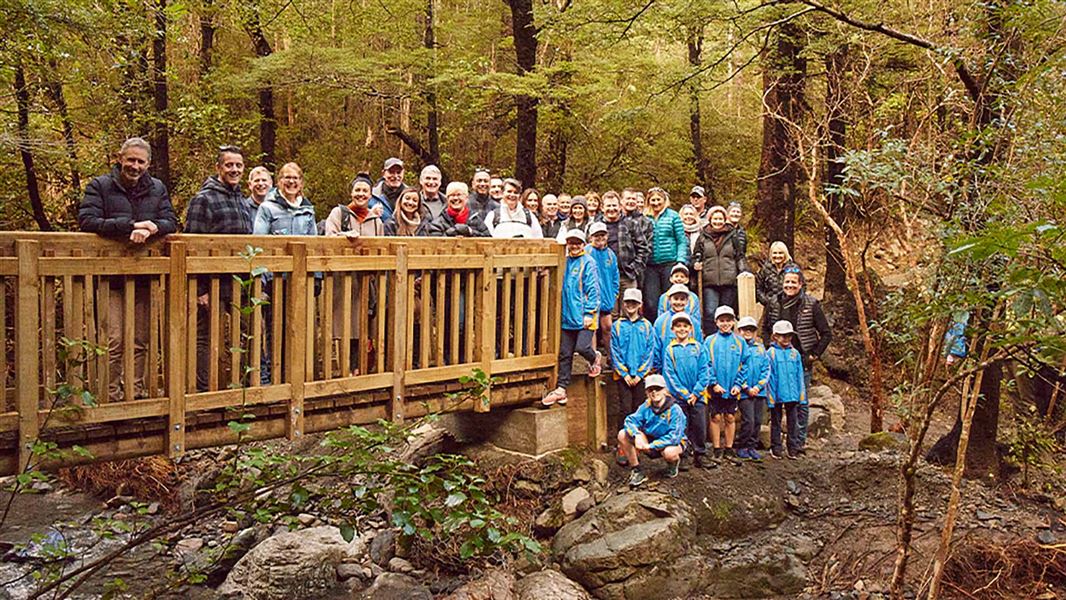 Group of people stand on and beside a bridge over a stream in the forest.