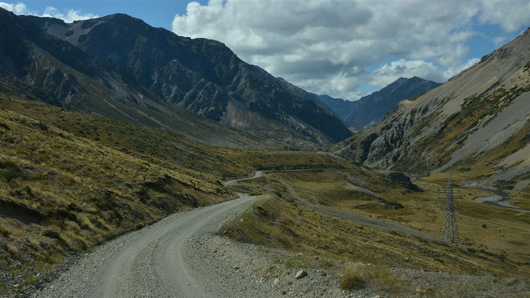 A gravel road winds through a steep rugged mountain valley, followed by power pylons..