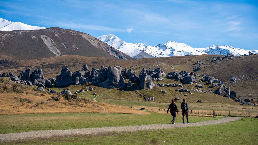 Two walkers follow a track through a landscape with golden tussock, limestone rock formations and snowy mountains.