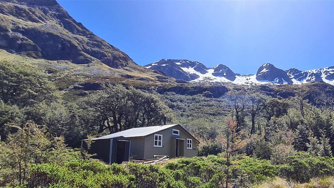 Green weatherboard hut in trees with snow capped ridge behind.