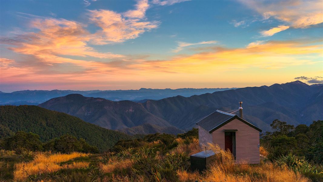 A view of a small hut in the foregrand with the forested ridges of Kahurangi Forest Park stretching into the distance.