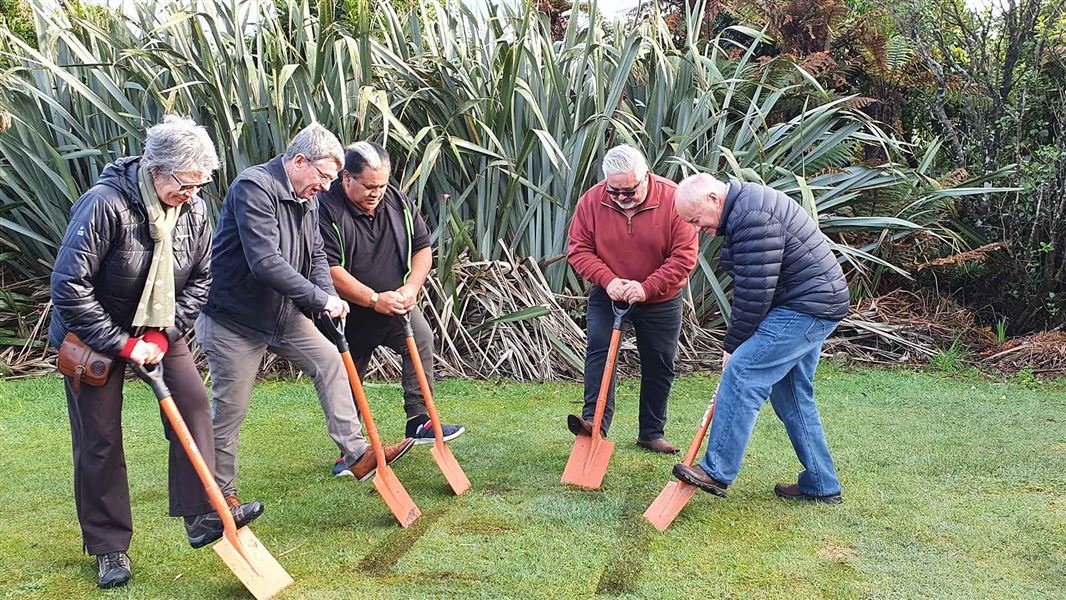 Five people are about to use a shovel to turn up the grassy ground.