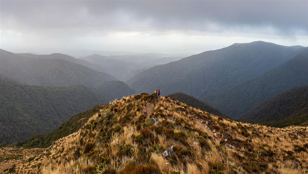 A distant view of two people atop a hill, face a rolling range of mountains.