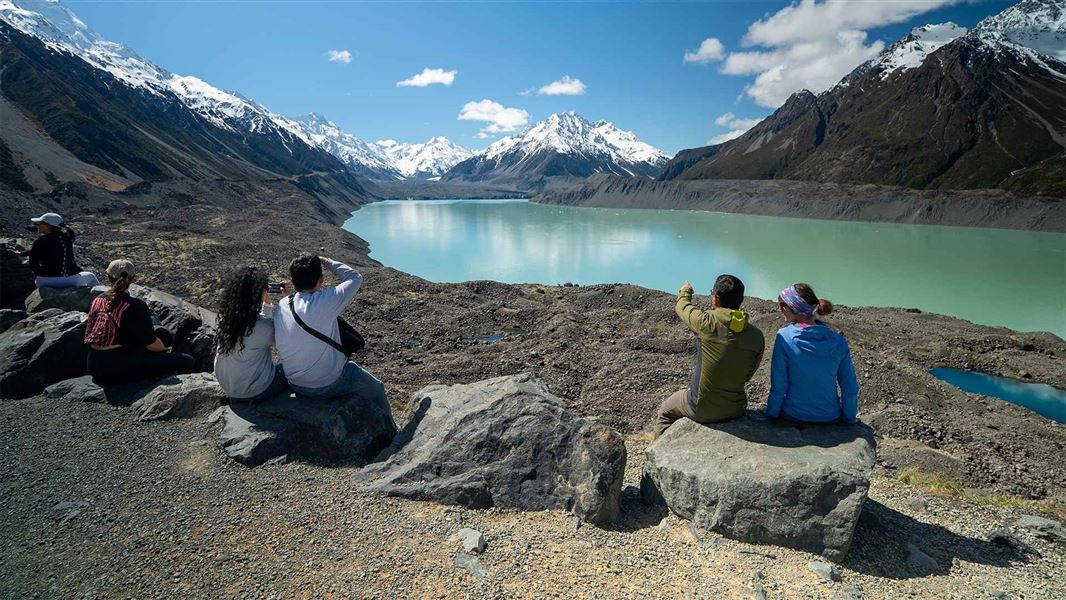 Groups of walkers sit on rocks overlooking a turquoise glacial lake ringed by snow-capped mountains.