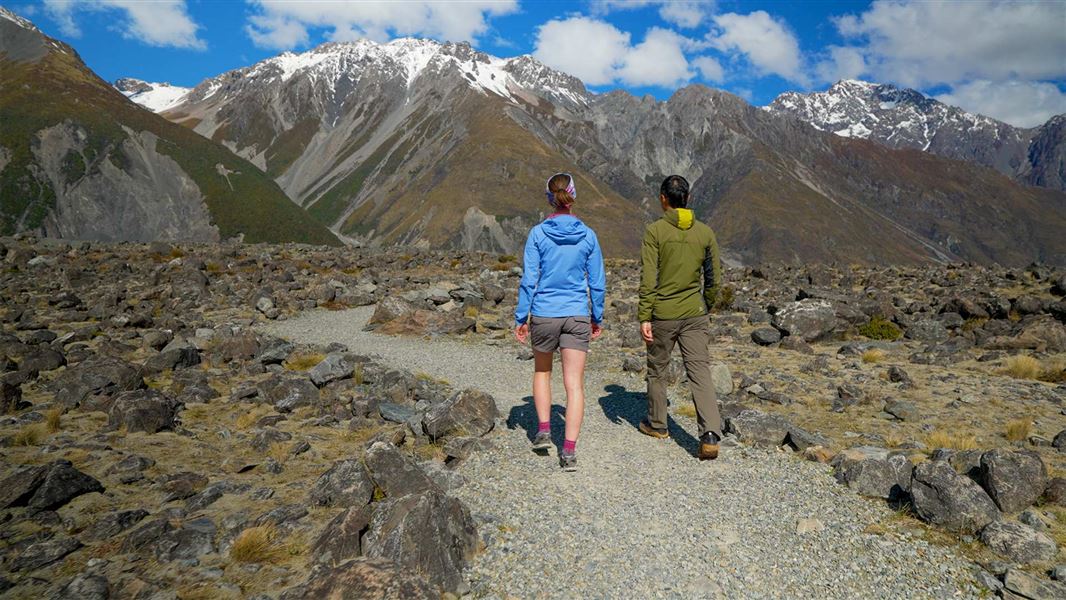 Two walkers follow a gravel track through rocky glacial debris toward jagged mountains.