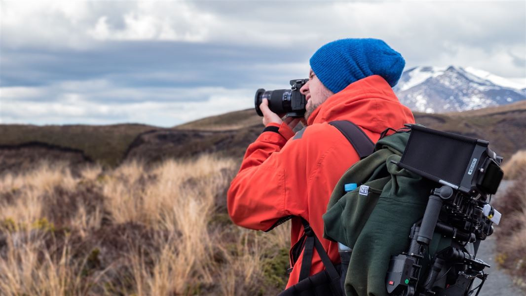 A man stands in an orange coat taking photographs with a professional but compact camera.