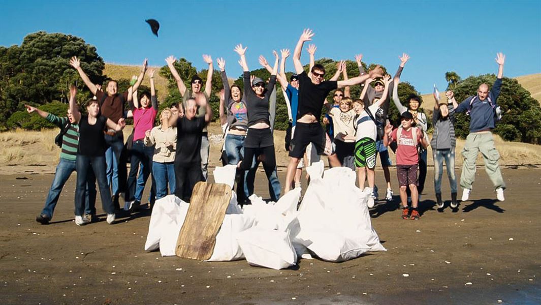 A group of people jumping together on a beach, behind several full bags of trash. 