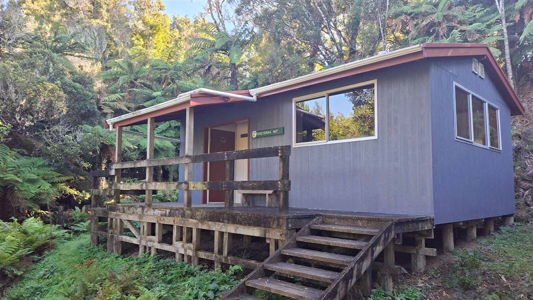 A small gray hut with aluminium window frames and a wooden deck with stairs sits nestled in the bush. 