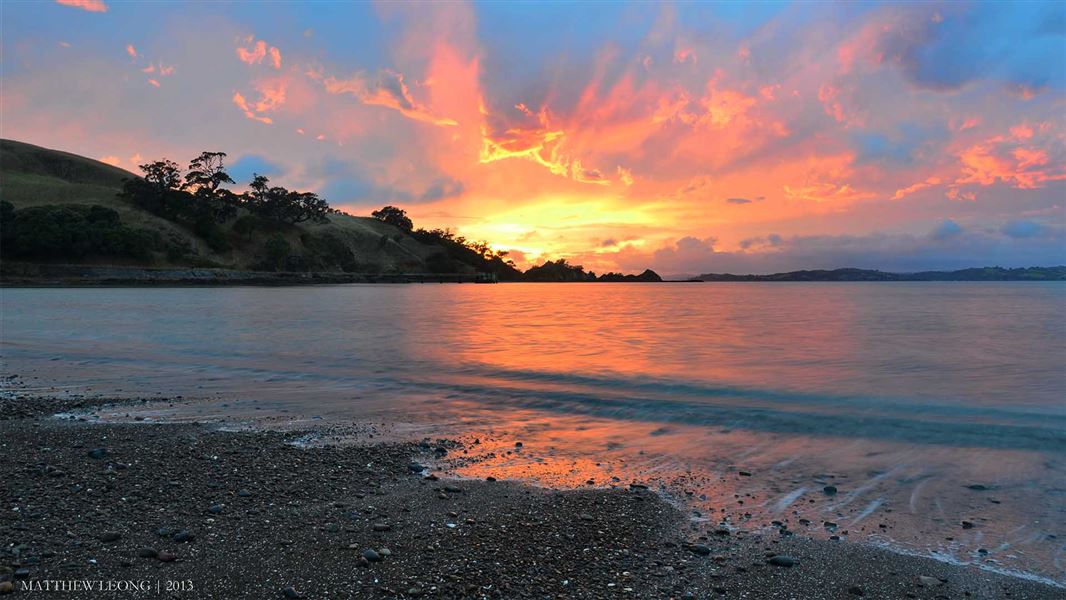 A bright sunset descends behind some hills in the distance. There is a large bay before the hills. The strong colours of the sunset reflect in the water.