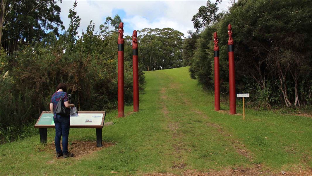 A person stands before an information panel. Four tall red coloured Māori totems mark the entrance to an open area.