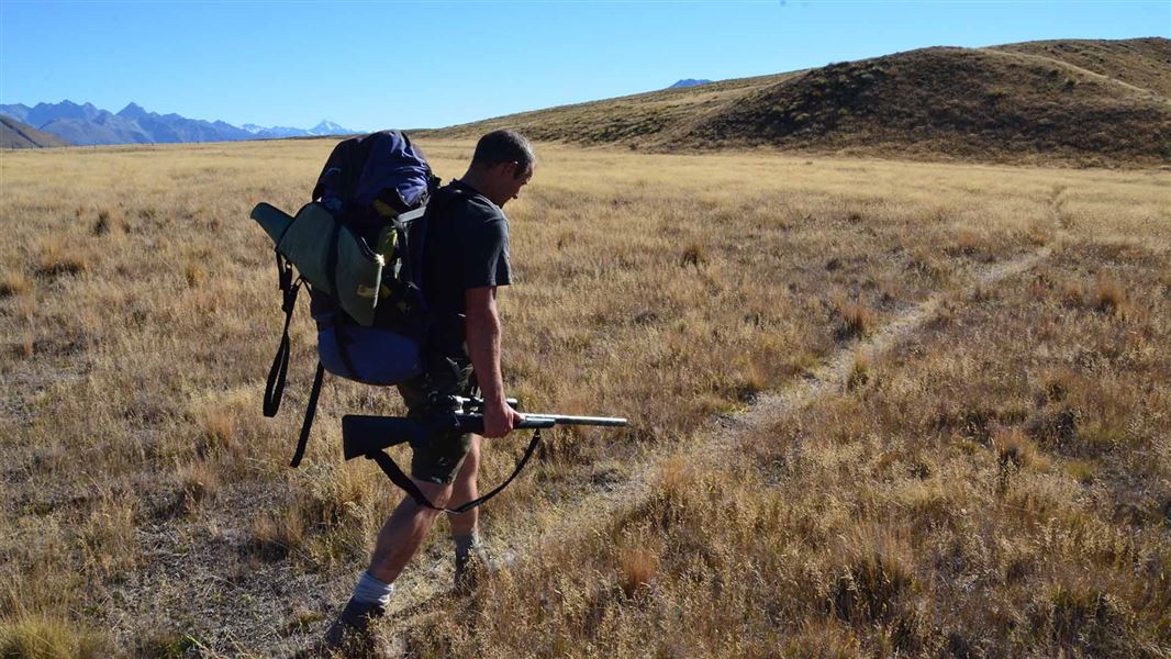 A person with a pack and a rifle walks across a plain covered in grass. There are small hills in the distance