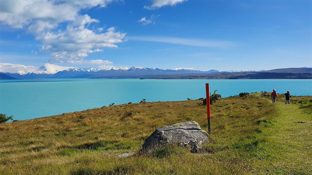 Children walk along a grassy hill above a turquoise lake, mountains in the distance.