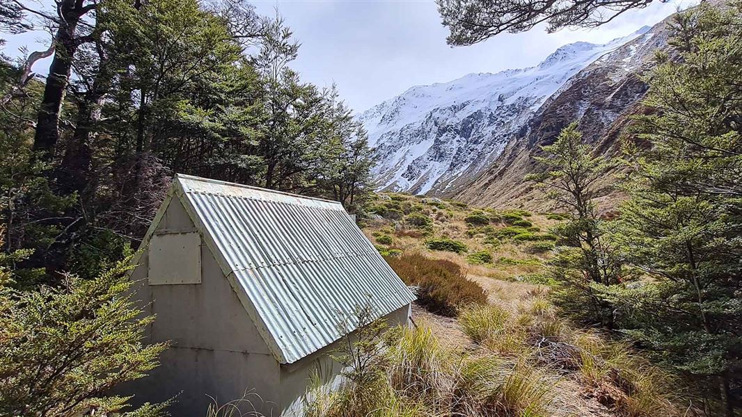 A tin-roofed hut surrounded by beech forest with a view of snow-capped mountains in the distance.