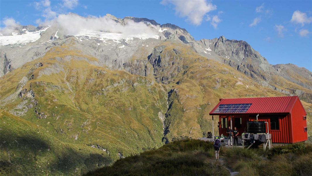 People in tramping gear approaching a hut on a hill overlooking a mountain.