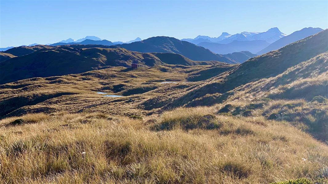 Views from atop a small grassy hill. Other hills covered in grass continue for a long distance before transitioning to large mountains.