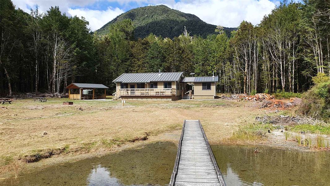 A one story tall wooden hut in an opening next to a forest. The hut also sits on the edge of a lake.